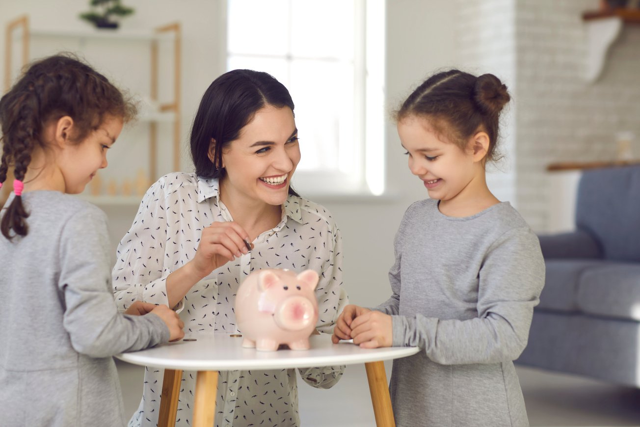 Mom teaches her two little daughters financial literacy and puts money in a piggy bank.